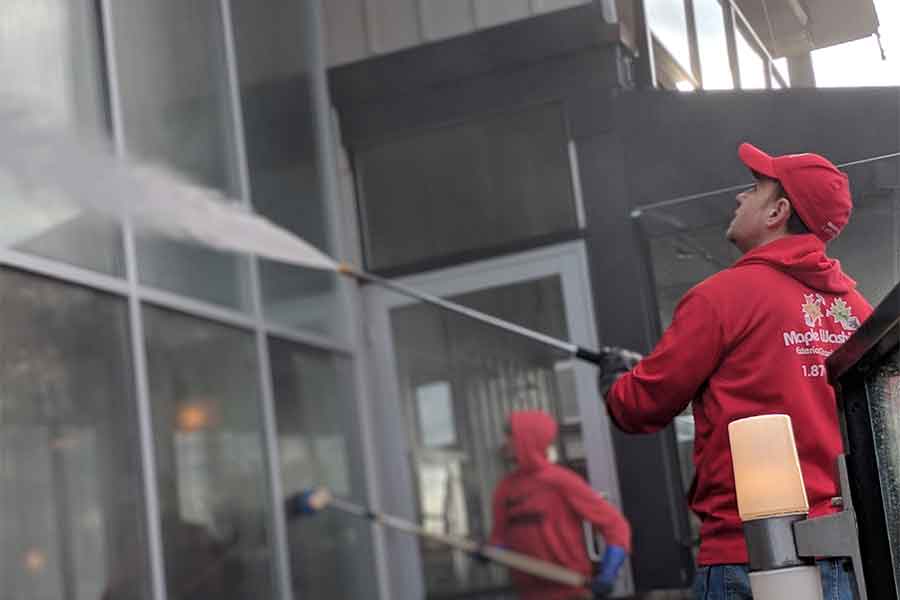 Two Maple Washing technicians in red uniforms use high-pressure washers to clean a large glass exterior, removing dirt and grime from building surfaces during a professional power washing service.