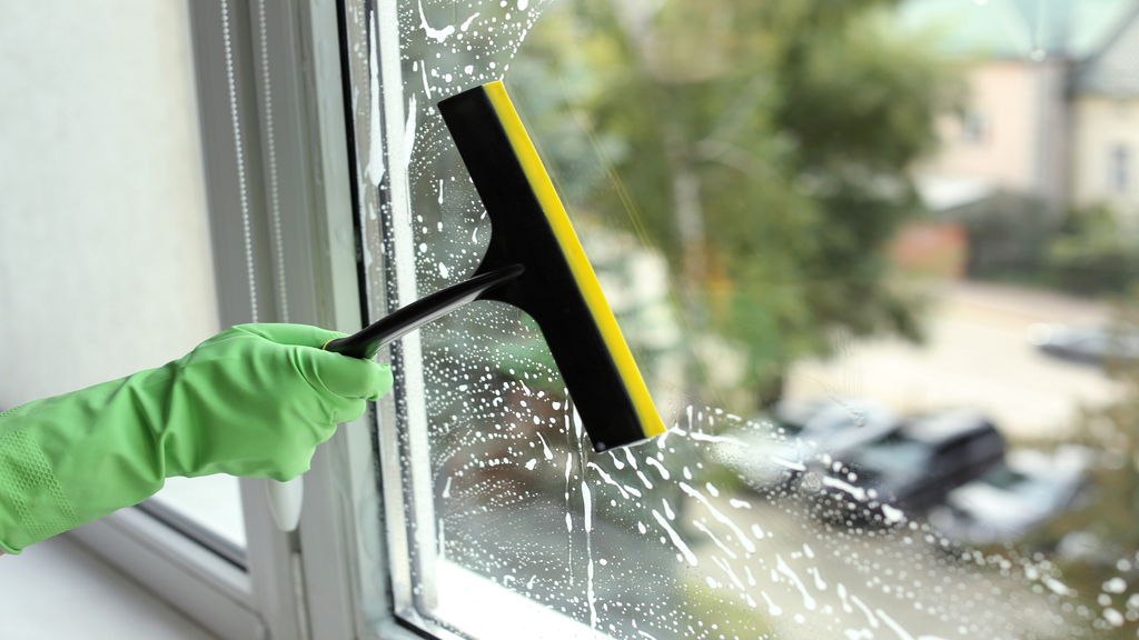 The Difference Between Hand-Washed Windows and Pole Systems1 Close-up of a person cleaning a window by hand with a squeegee, wearing a green glove, removing soap for a streak-free finish.